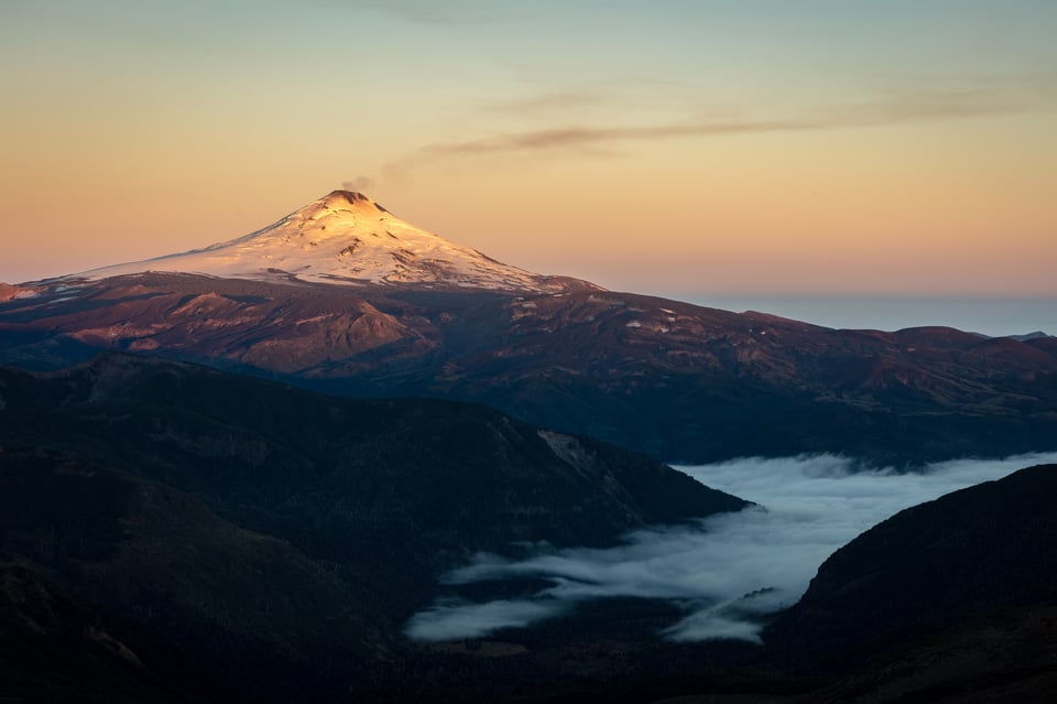 The Volcan Villarica Volcano is visible from a nearby mountain at sunrise, with thin plumes of smoke rising into the sky.